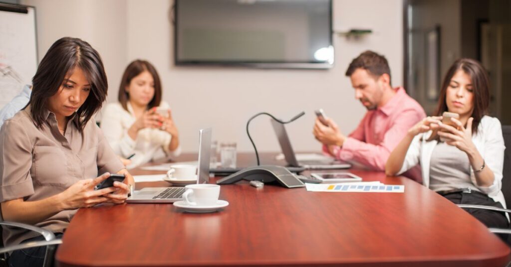 A team seated around a conference table, each person focused on their own device, highlighting the struggle of leading in a digitally distracted workplace.