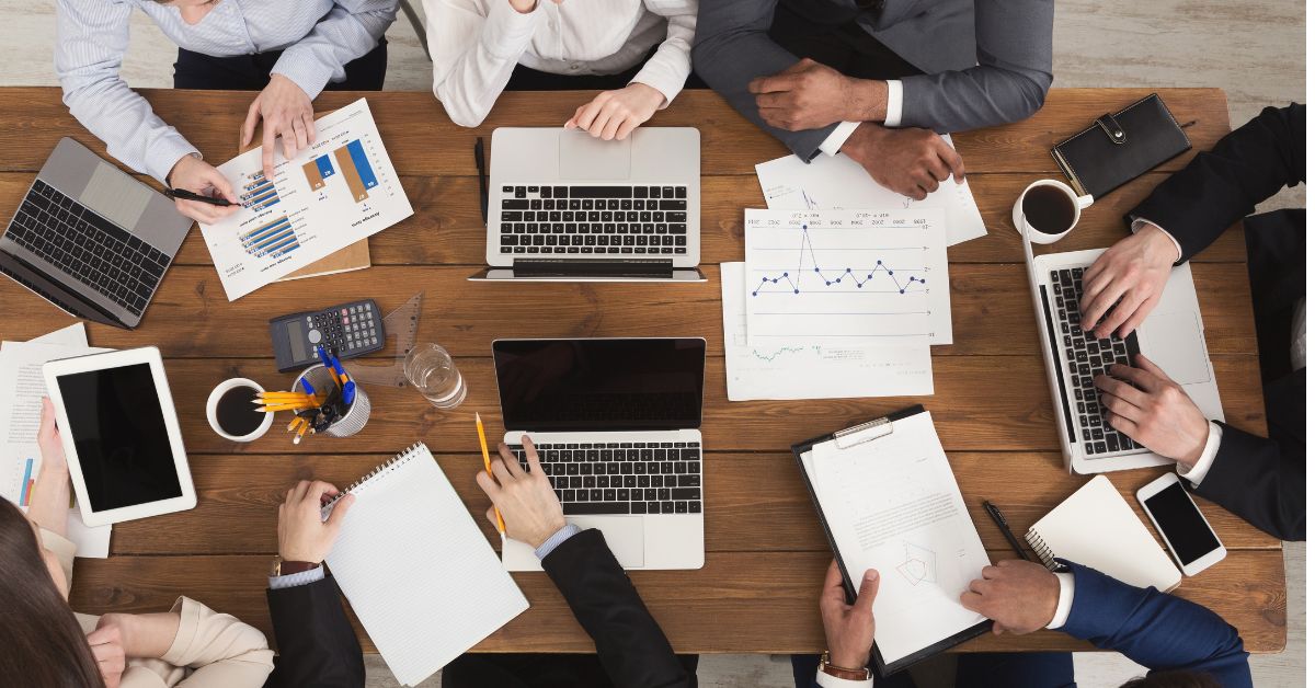 A group of people sitting around a meeting table, each distracted by their devices illustrating the challenge of leading a digitally distracted team.