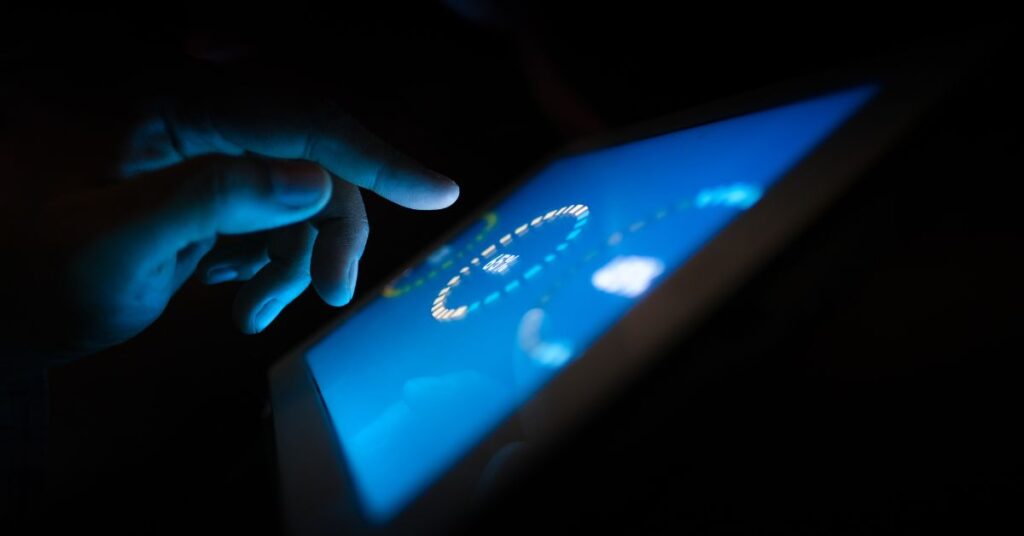 A man's hand tapping an ipad screen that is shining with blue light in the darkness
