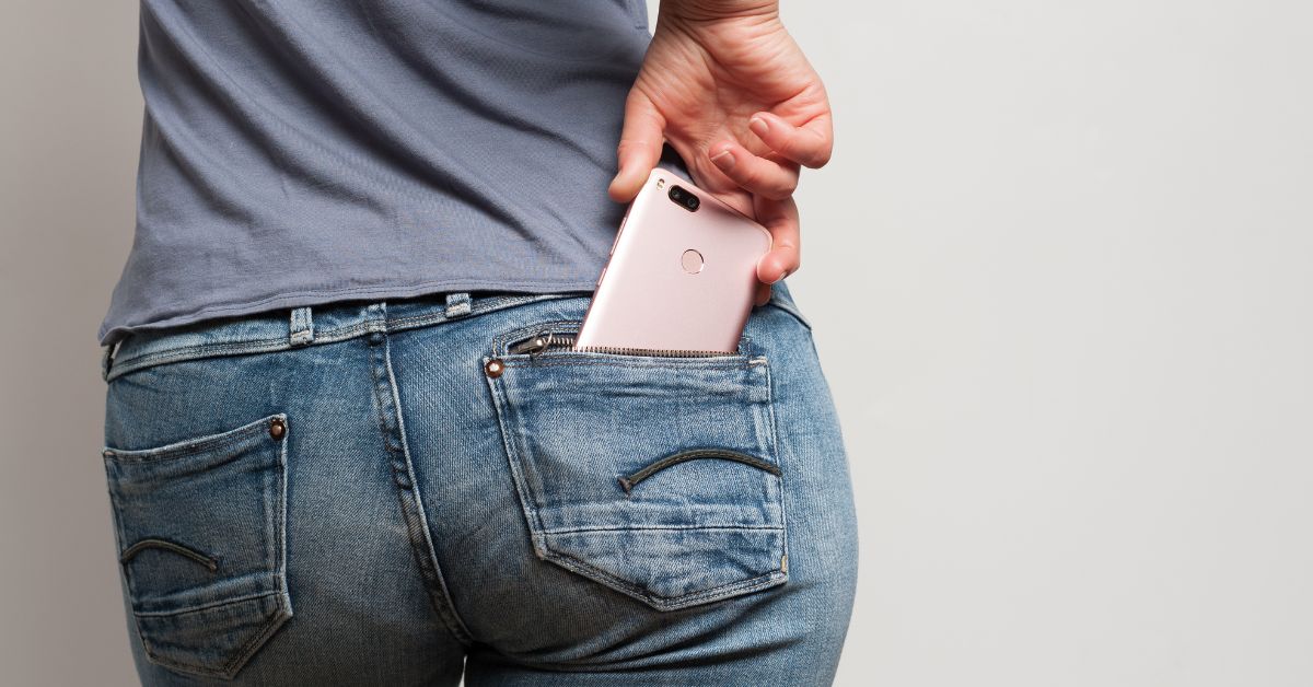 A woman standing with her back facing you with a pink cell phone in her back jean pocket representing how attached we always are to our phones.