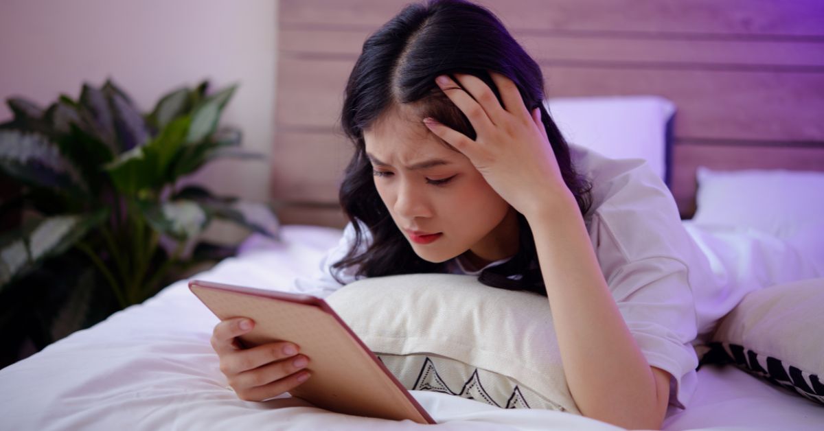 Asian woman lying on her bed with an ipad holding her head in distress from doomscrolling.