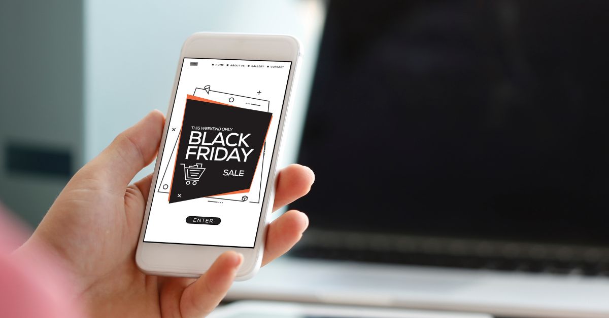 A woman's handing holding a cell phone with the words Black Friday on the screen and a large computer screen in the background.