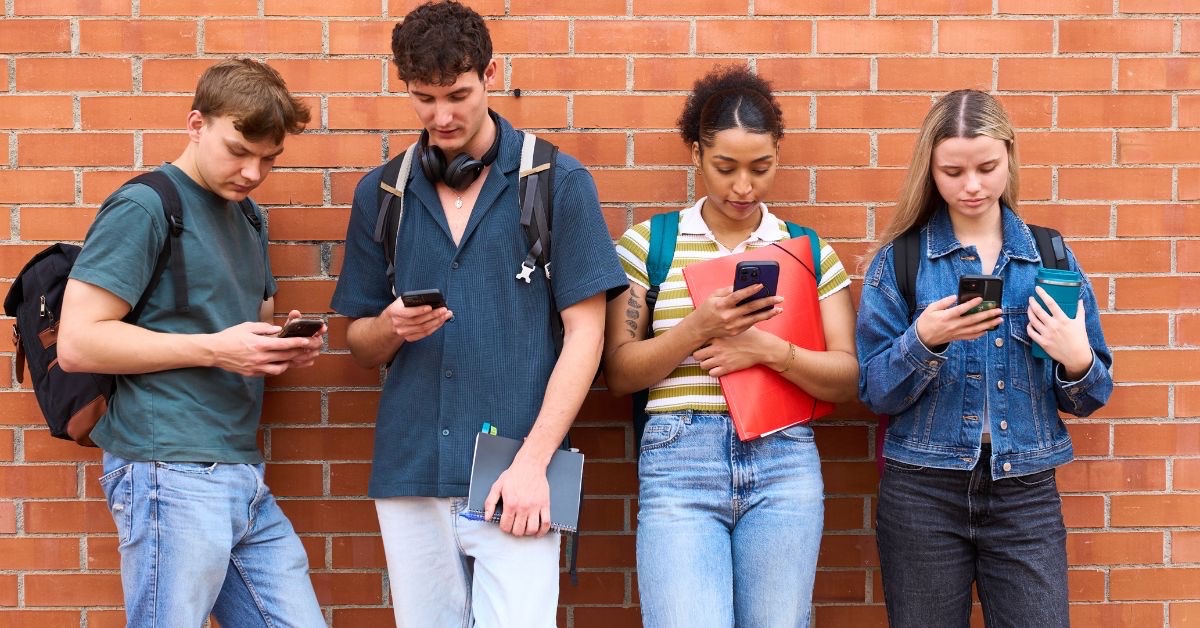 4 unfocused young adults with school books and knapsacks leaning against a brick wall all looking at their phones having lost their attention to their phones.
