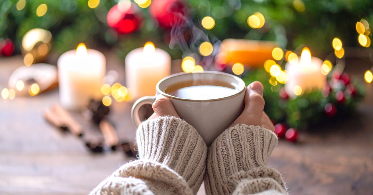 A person in a cozy knit sweater holding a steaming mug of coffee or tea in front of glowing candles and holiday lights, evoking a peaceful moment to log off and unwind.