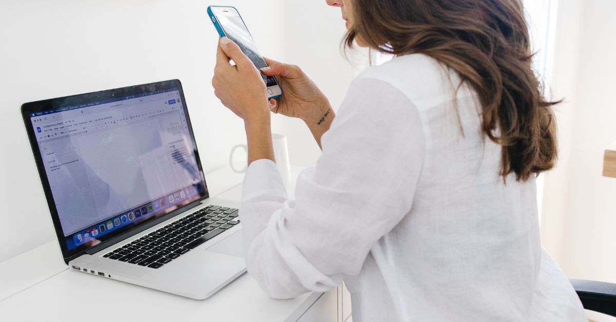 Woman in a white shirt sits at a desk with an open laptop, focusing intently on her smartphone while ignoring the screen in front of her, illustrating phone addiction. The image captures distraction and over-reliance on mobile devices, even during productive tasks.