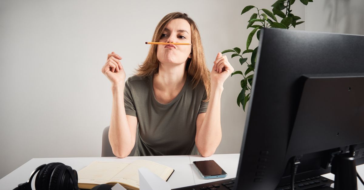 Women in front a a computer screen on a desk with a phone in front of her as well. She is killing time balancing a pencil between her lips and nose. It is obvious she is procrastinating.