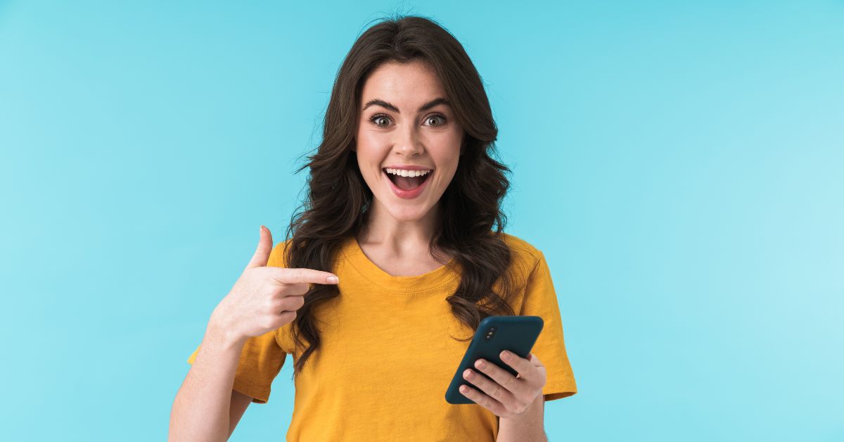 woman in a gold shirt pointing at her phone with a very big smile on her face.