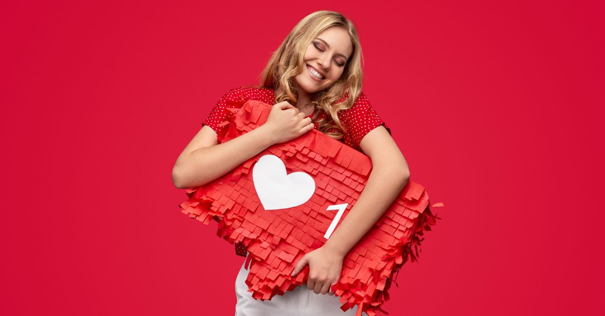 Young woman in a red polka dot top smiles while hugging a large red piñata shaped like a social media notification with a white heart and the number one, symbolizing social media addiction. The vibrant red background and her joyful expression highlight emotional attachment to online validation.