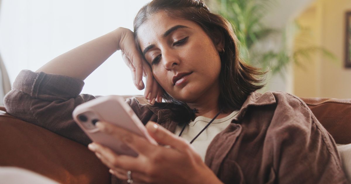 Young woman lounges on a couch with her head resting on her hand, staring blankly at her phone in a disengaged, tired posture. The image captures the emotional toll of endless scrolling, illustrating the need to learn how to stop doomscrolling.