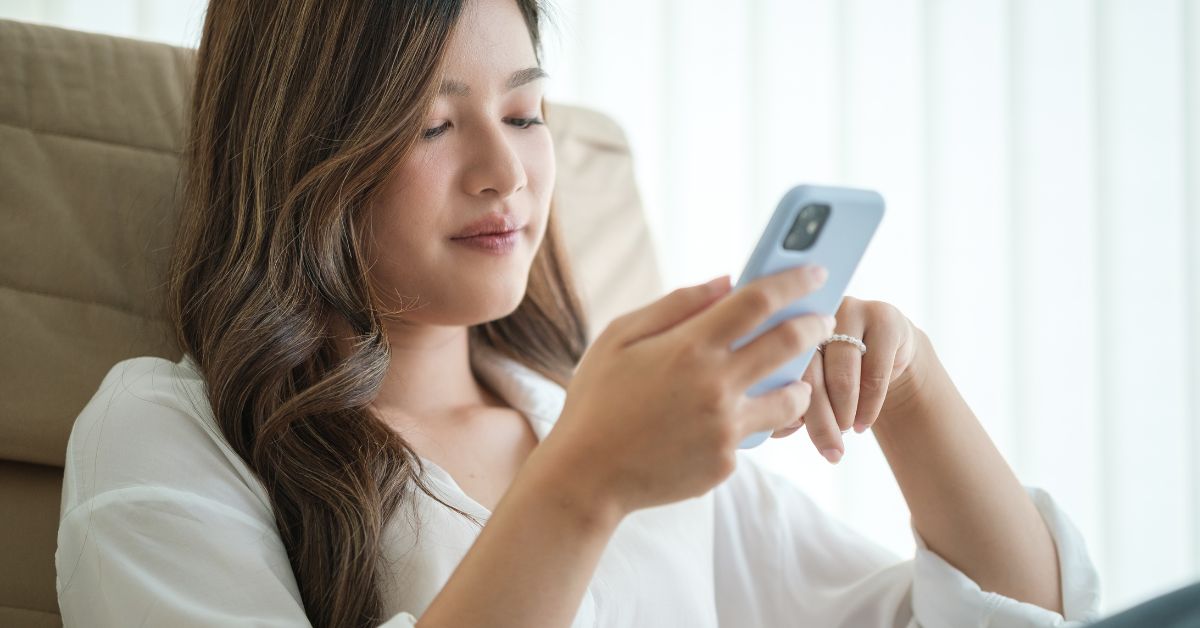 Asian woman sitting smiling scrolling on her light blue phone.