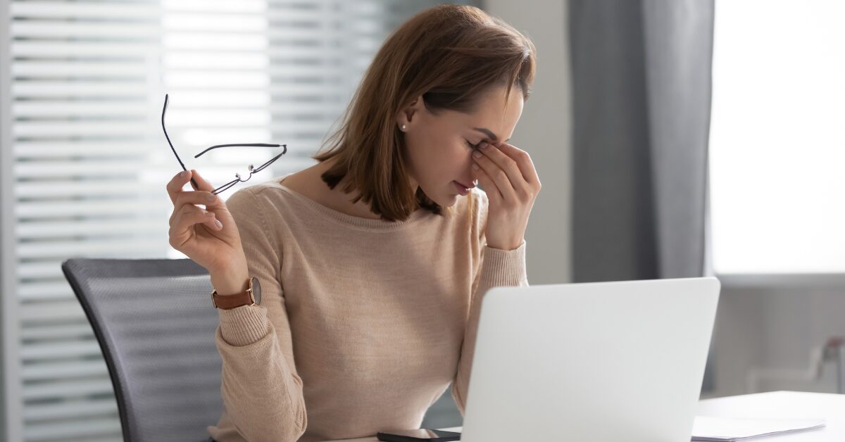 Woman sitting at a desk holding her glasses and rubbing her eyes in discomfort while looking at a laptop, showing signs of digital eye strain.