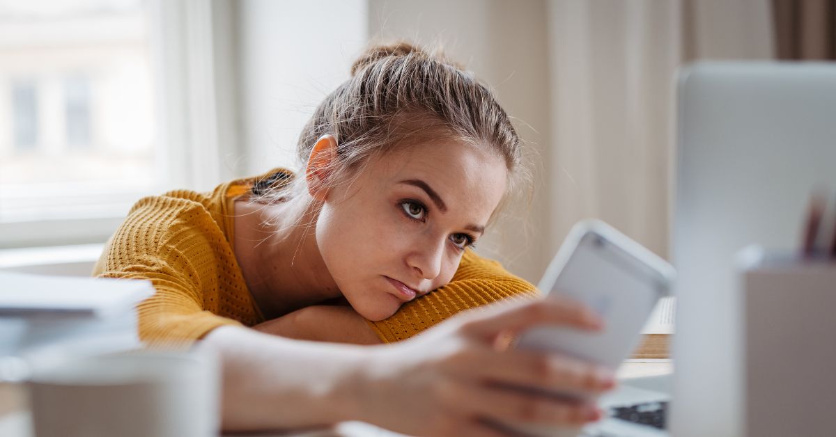 Woman in a mustard yellow sweater resting her head on her arm while looking at her smartphone beside a laptop on a desk. Soft natural light from a nearby window highlights her tired expression, suggesting distraction or digital fatigue while working from home.