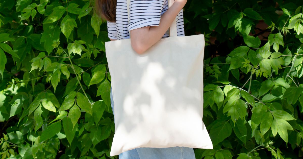 Person wearing a striped shirt holds a plain canvas tote bag against a backdrop of green leaves, illustrating a simple analog bag that can be filled with items to reduce digital distraction and break phone habits.