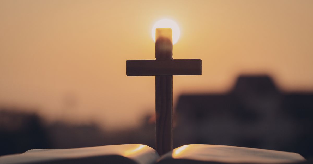 Wooden cross standing on an open Bible with the sun rising directly behind it creating a glowing halo effect. The peaceful sunrise scene reflects themes of hope and reflection often associated with an Easter devotional.
