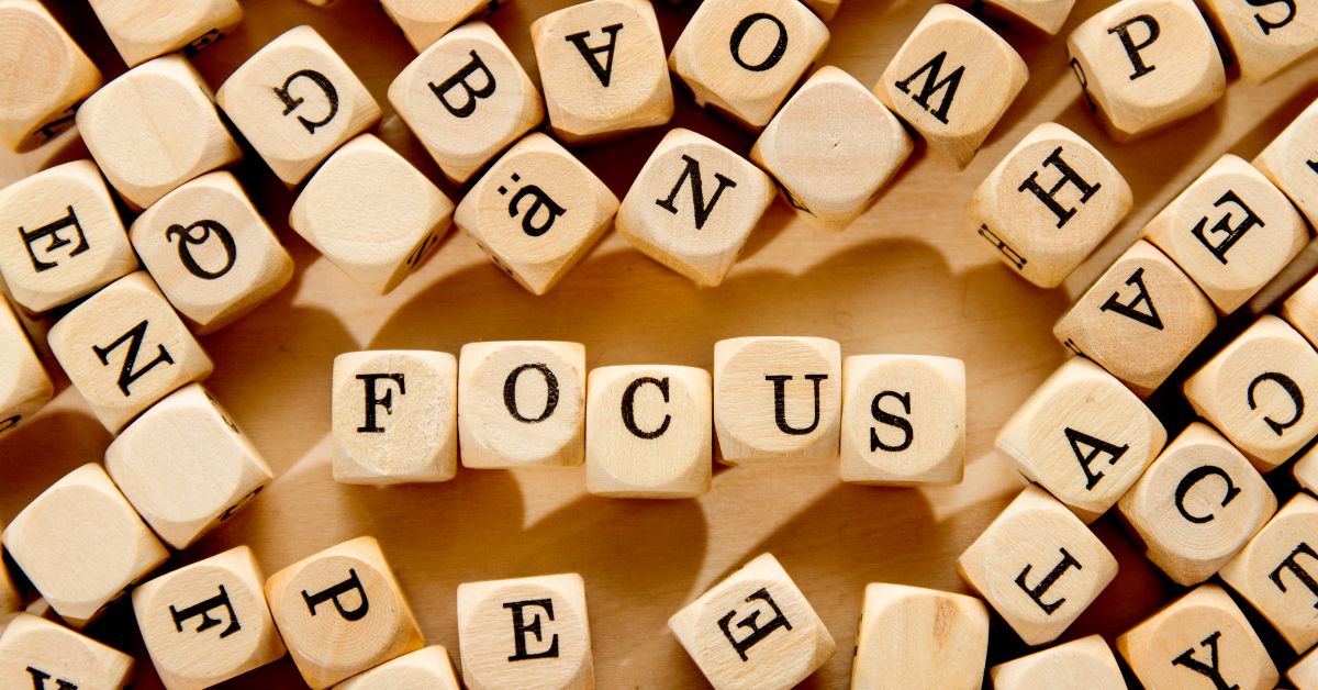 Close up of wooden letter tiles scattered across a light wood surface with the word FOCUS spelled clearly in the center. The surrounding tiles display random letters, drawing attention to the central word and visually emphasizing the concept of concentration and clarity. The warm tones and soft shadows create a calm, motivational atmosphere.