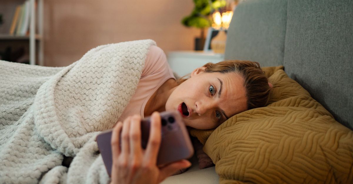 Woman lying on a couch under a blanket staring at her phone with a tired and absorbed expression late at night, illustrating doomscrolling and its impact on rest and focus. The scene reflects how to stop doomscrolling by highlighting excessive screen use in a relaxed home setting.