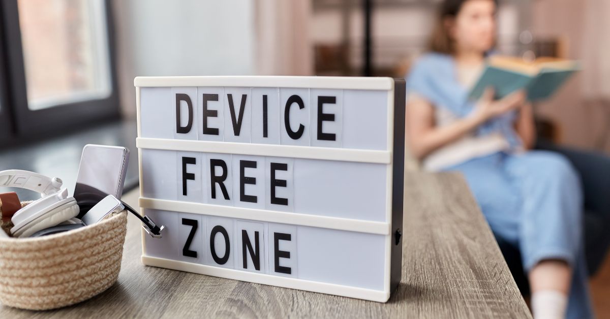 Lightbox sign on a wooden table reads "DEVICE FREE ZONE". A basket beside it holds a smartphone headphones and a charging cable while a woman sits blurred in the background reading a book.