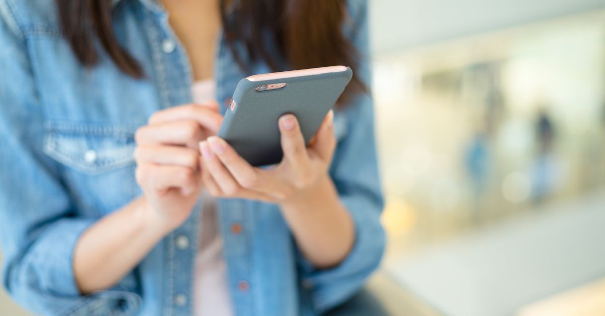Person in a denim shirt looking down while scrolling on a smartphone with both hands in a softly blurred indoor setting, representing mindless scrolling on a mobile device.
