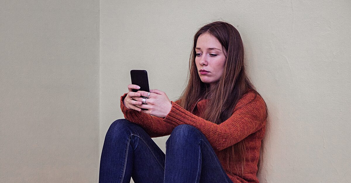 Young woman in a rust-colored sweater sits on the floor against a plain wall, staring at her phone with a tense, withdrawn expression. Her body is curled inward with knees pulled up, reflecting phone anxiety and the isolating pull of scrolling for comfort.