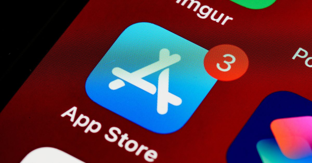 Close up of the Apple App Store icon on a phone screen with a red notification badge showing "3". The word "App Store" appears below the blue icon on a red app folder background.
