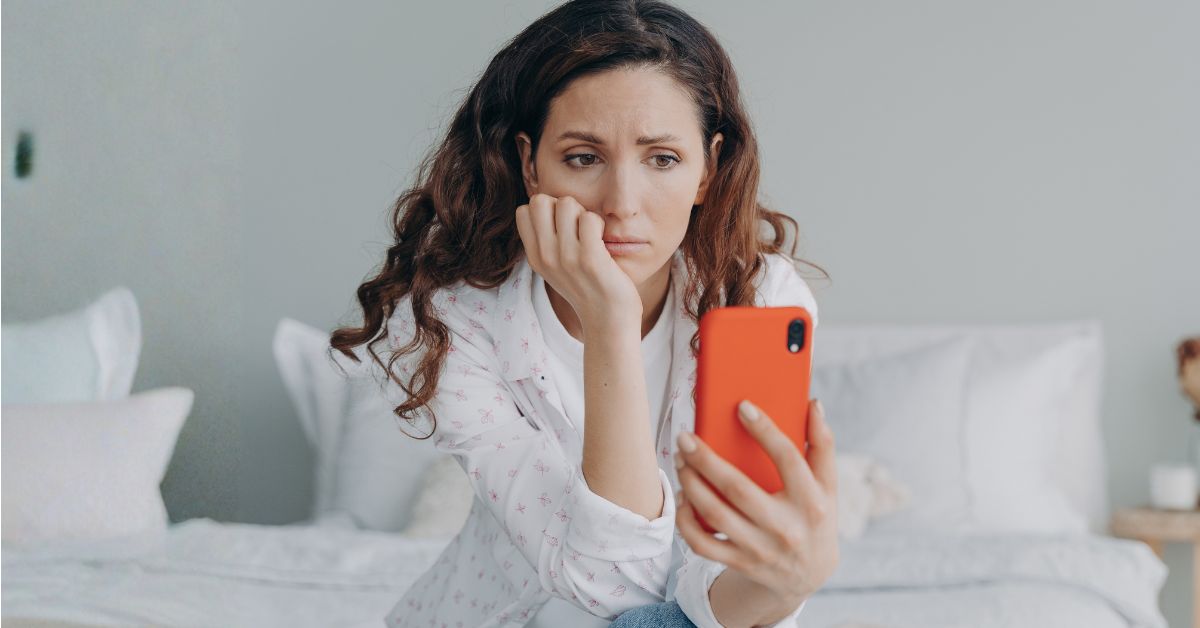 A worried woman sits on a white bed while looking at an orange phone. Her tense expression and slumped posture suggest the fear and anxiety that can come from doom scrolling.