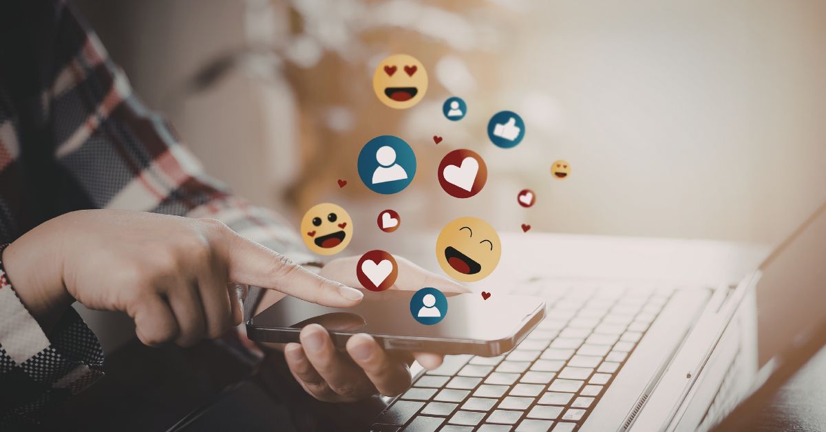 A person is checking social media on a smartphone beside an open laptop. Floating reaction icons with hearts likes and smiling faces show online engagement coming from the phone.