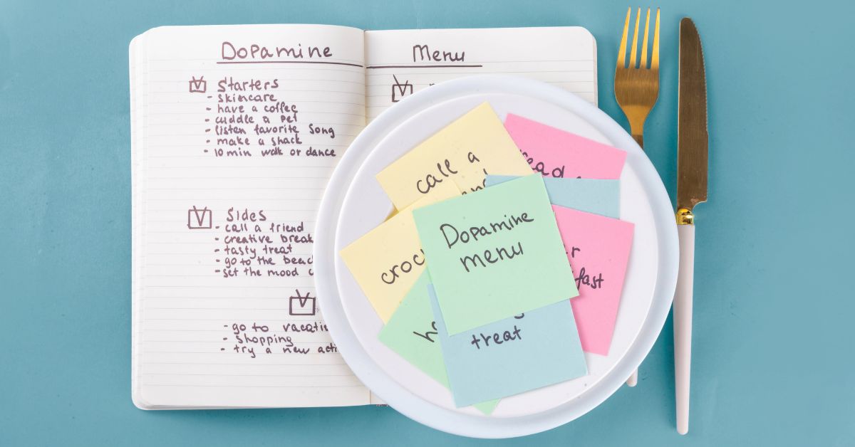 Open notebook titled “Dopamine Menu” beside a plate of colorful sticky notes, with a gold fork and knife on a blue background. The notes include ideas like “Dopamine menu” and “treat” for planning mood boosting activities.