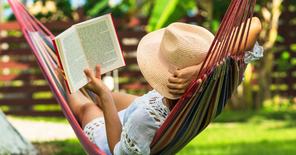 A person in a straw hat rests in a striped hammock outdoors while reading a book in a sunny garden. The peaceful setting connects spiritual rest with quiet time away from daily distractions.