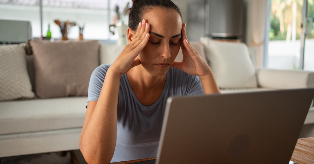A woman sits at a laptop with her eyes closed and both hands pressed to her temples. Her tense posture suggests brain fog and discomfort after prolonged screen time.