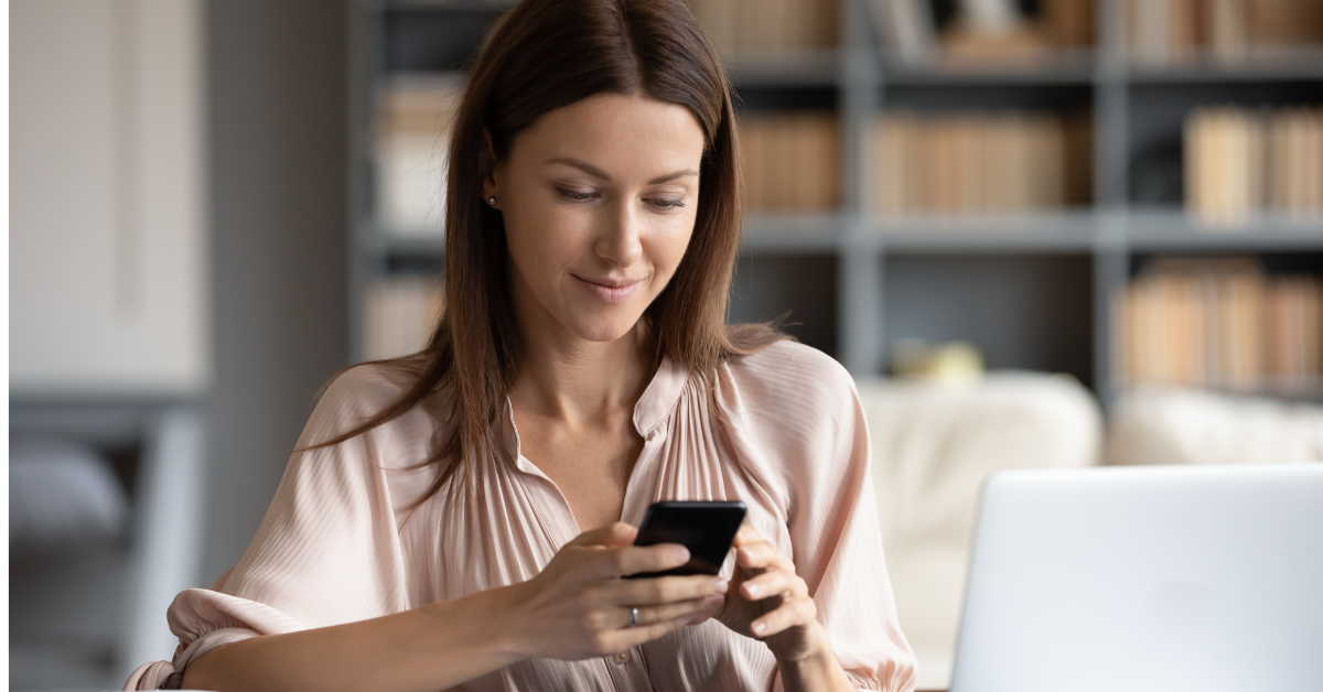 A woman checks her phone beside an open laptop in a bright home office. The scene highlights digital boundaries around when and how quickly to reply to messages.