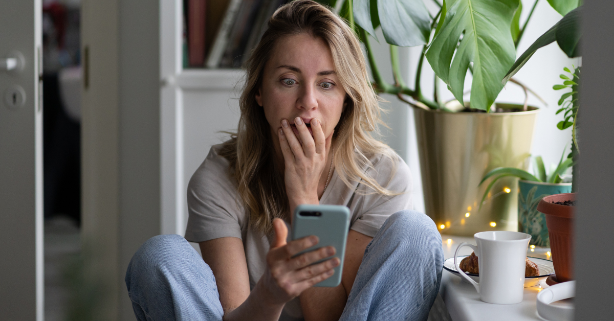 A woman sits on the floor by houseplants and a coffee mug staring wide eyed at her phone with one hand over her mouth. Her anxious expression suggests the overwhelm of a messy phone screen and supports a post about digital declutter.