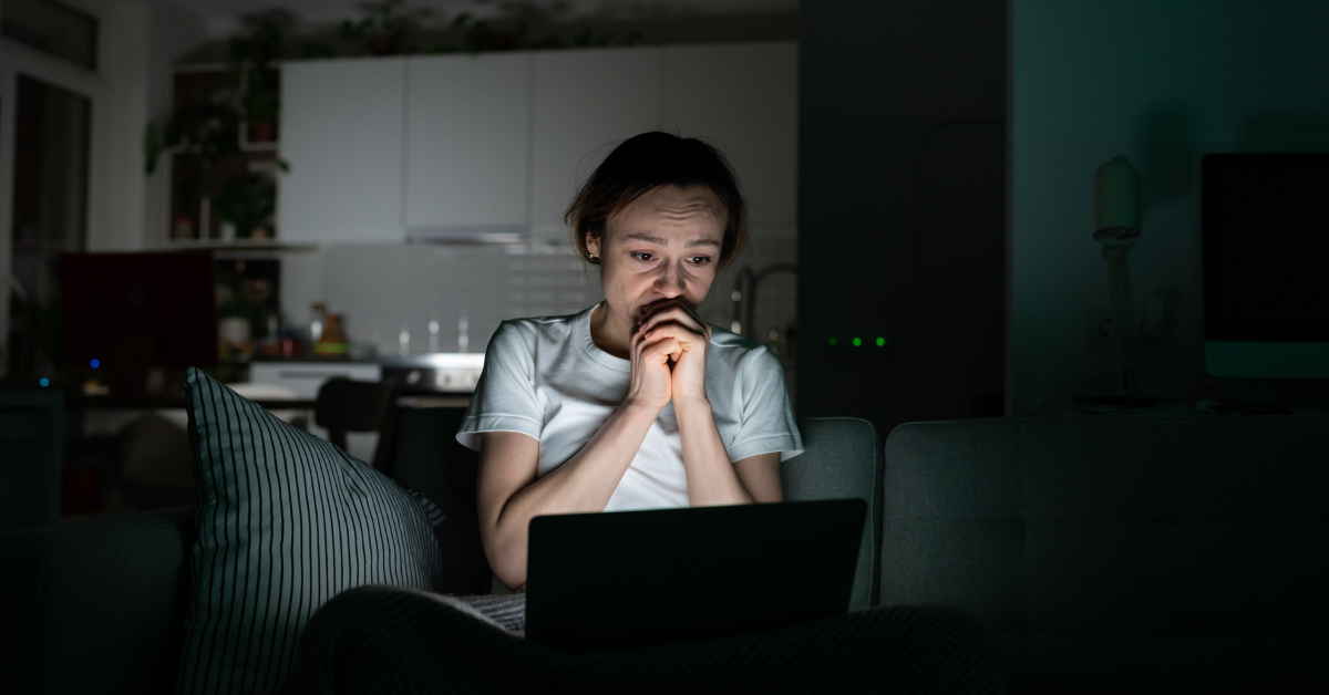A worried person sits on a couch in a dark room with their hands clasped while staring at a glowing laptop. The late night scene reinforces the reminder to stop scrolling before bed.