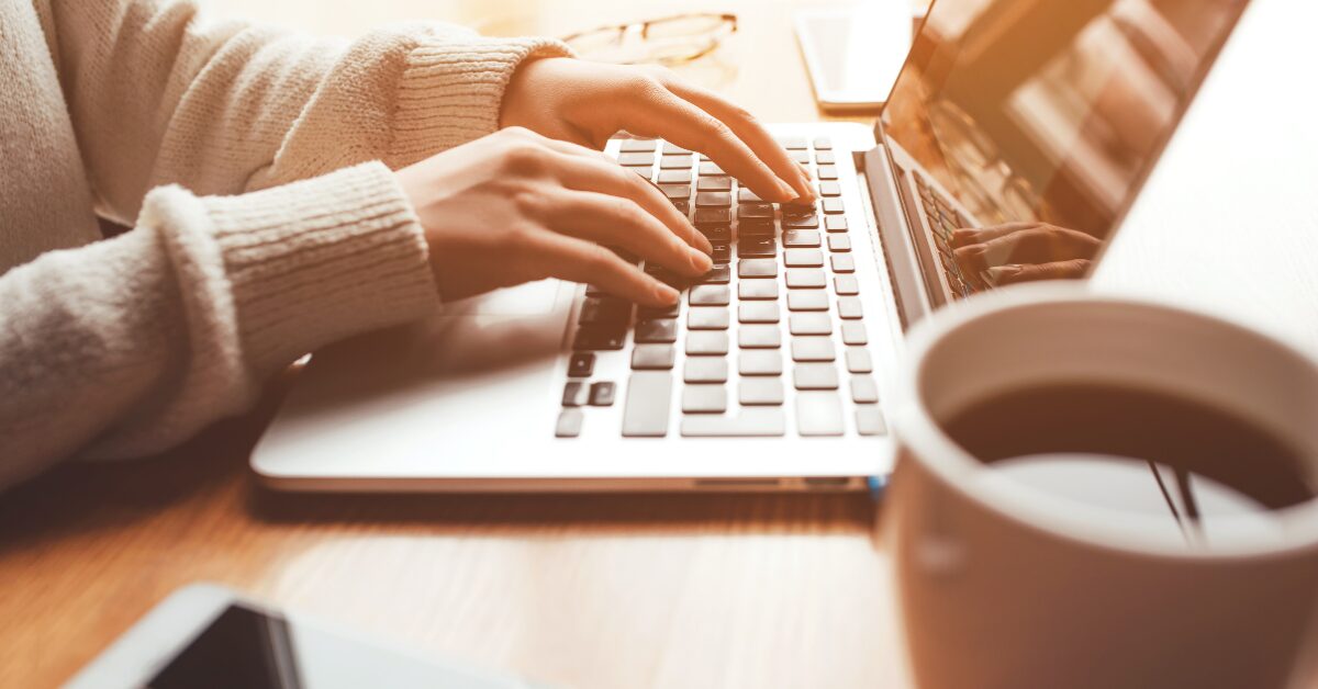 Person in a cozy sweater typing on a laptop beside a cup of coffee and a smartphone. The warm workspace setting illustrates everyday computer screen time.