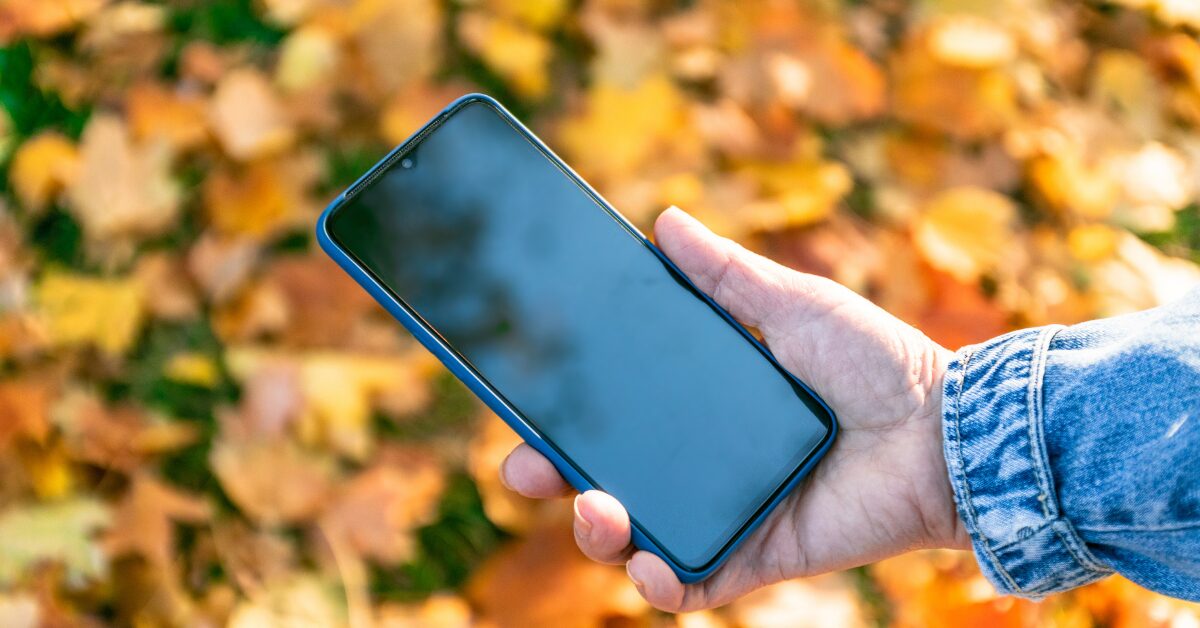 A person in a denim jacket holds a smartphone with a dark screen outdoors over a background of colorful autumn leaves. The scene represents everyday phone habits and how often people keep their devices close at hand.