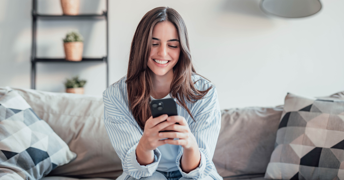 A woman sits on a couch smiling down at her phone in a bright living room with patterned pillows and small plants in the background. Her calm focused expression supports a post about how to declutter your phone so you can feel more peace stay focused and be more present in your relationships.