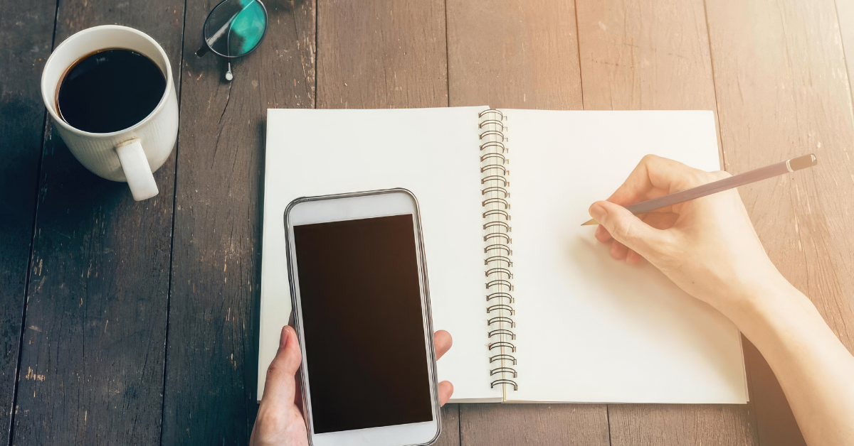 A person holds a smartphone beside an open blank notebook while writing with a pencil at a wooden table. The setup reflects writing out personal phone rules and deciding how to use a phone more intentionally.