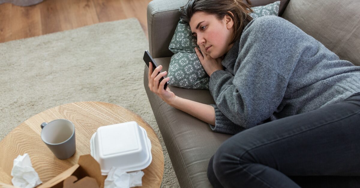 A weary woman wearing gray lies on her couch, staring at the phone beside takeout and crumpled tissues. This scene shows how doomscrolling news can quietly drain energy.