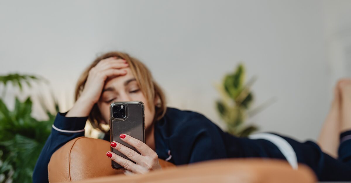 A weary woman in navy pajamas holds her phone while lying on the tan leather couch, palm pressed to forehead. This quiet moment gently reminds us scrolling is not restful.