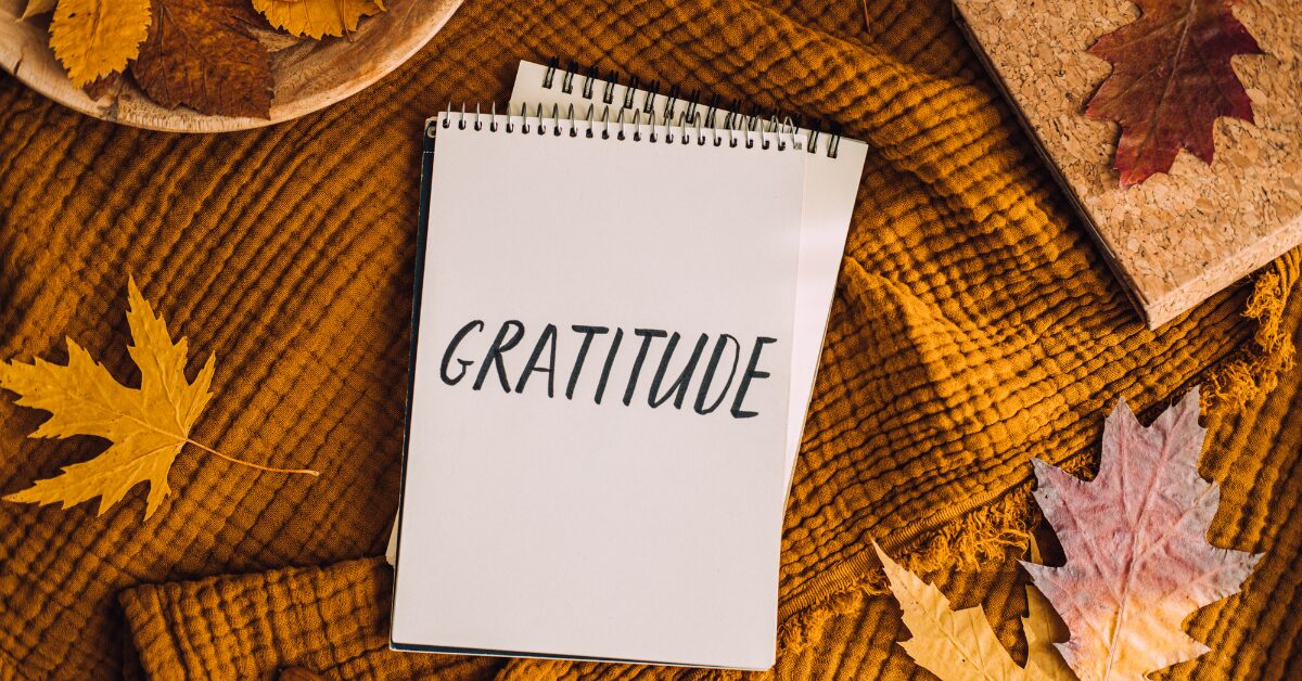 Spiral notebook with the word "GRATITUDE" surrounded by autumn leaves on a warm orange blanket. The scene represents how to practice gratitude using a simple gratitude practice you can do on the phone you already carry in your pocket.